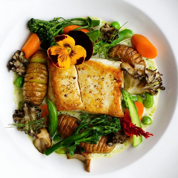 Pan-seared fish with hasselback potatoes, broccolini, carrots, mushrooms, and edible flower garnish.
