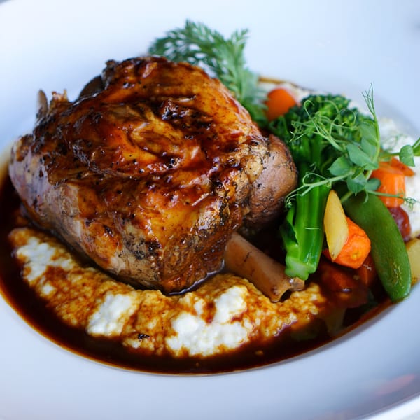 Braised lamb shank with gravy, polenta, and mixed vegetables on a white plate.