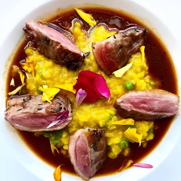 Seared duck breast slices on yellow risotto with brown sauce and colorful edible flower petals.