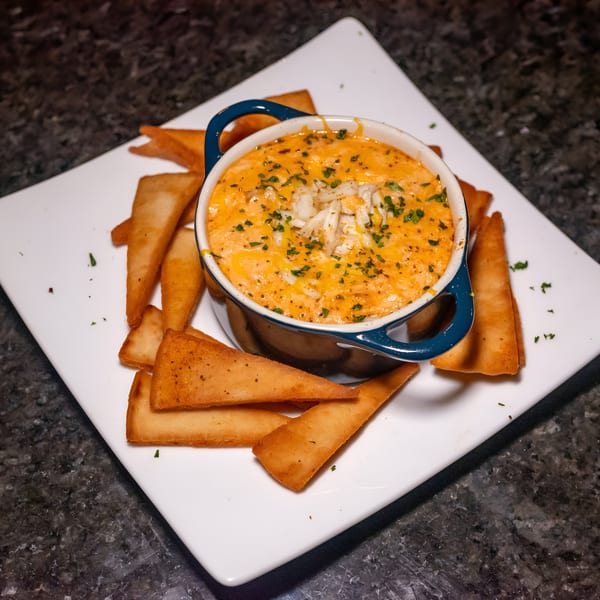 A blue ramekin of cheesy crab dip, garnished with parsley, surrounded by pita chips on a white plate.